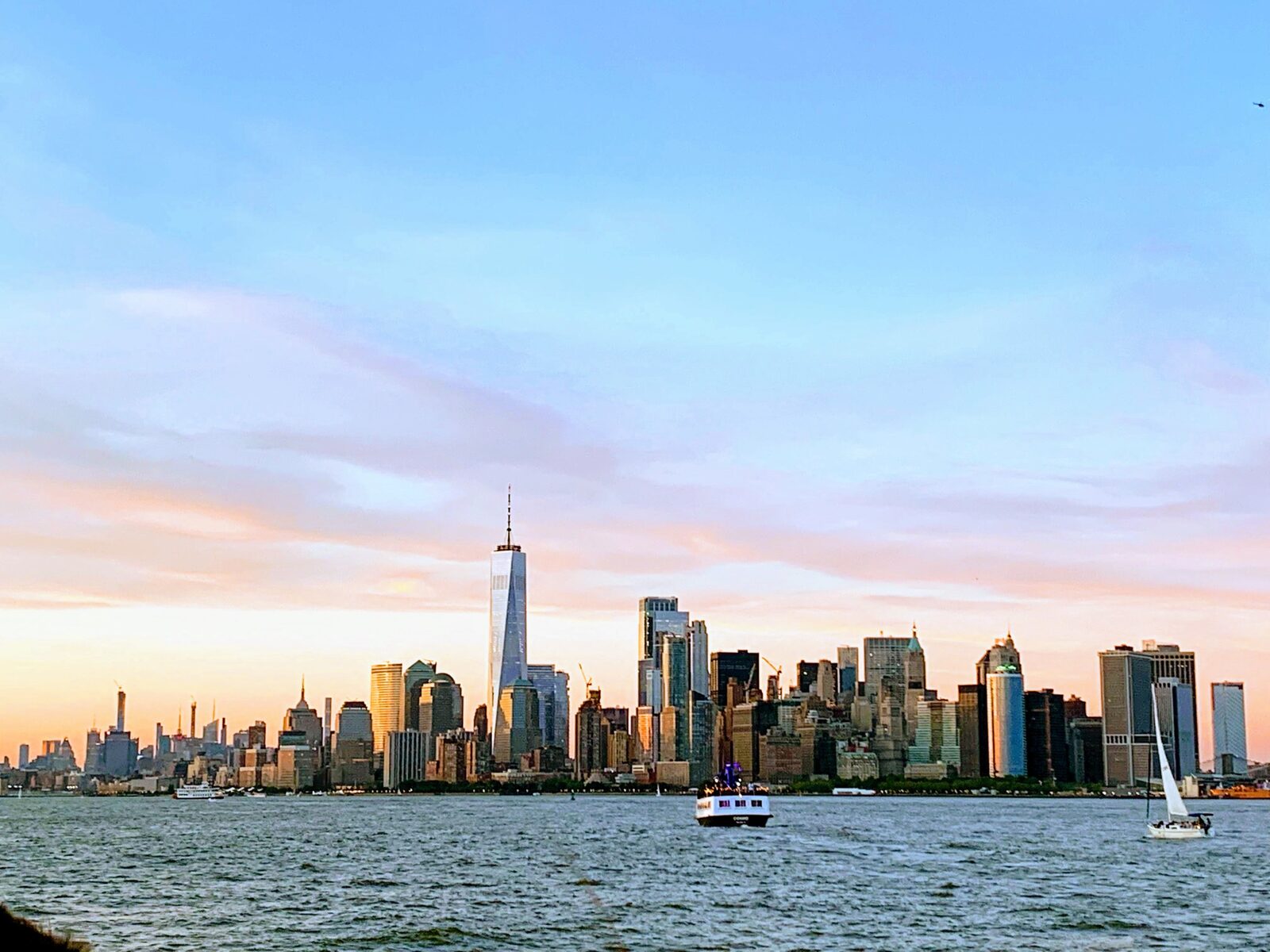 Lower Manhattan skyline across the harbor at sunset, a ferry and a sailboat on the water