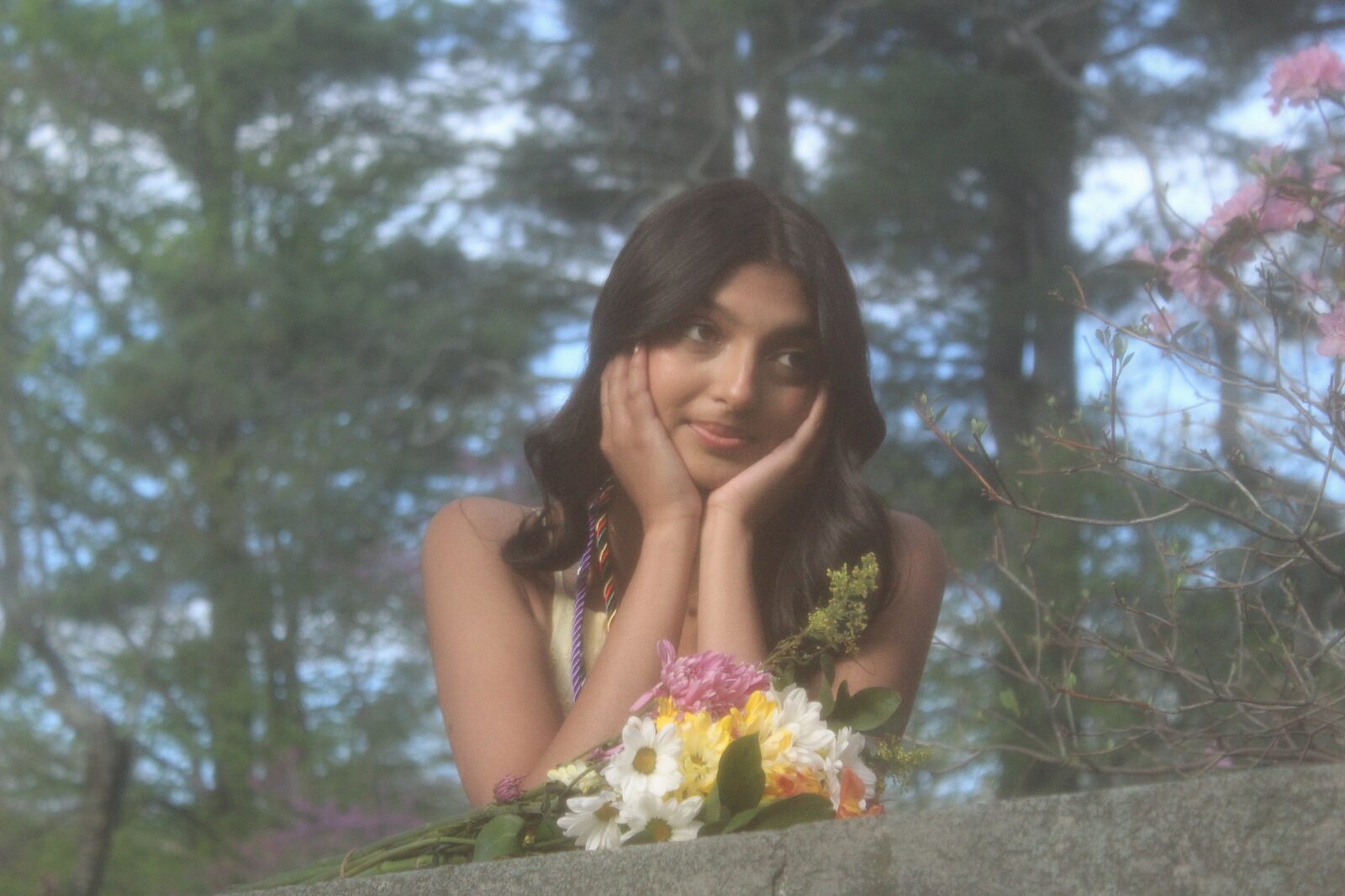 Kavya resting her face in her hands behind a stone wall, wildflowers and pine trees around her