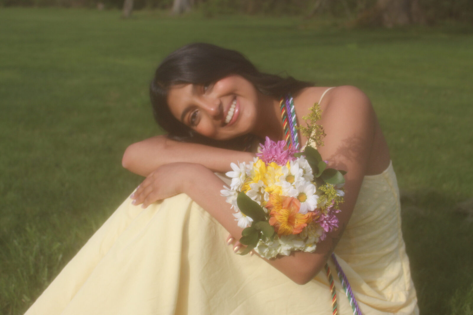 Kavya in a yellow dress smiling on the grass, her head resting on her arms, holding a bouquet